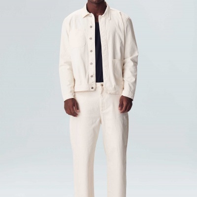 Homem com casaco e calças creme e ténis, t-shirt preta por baixo, fundo cinzento claro