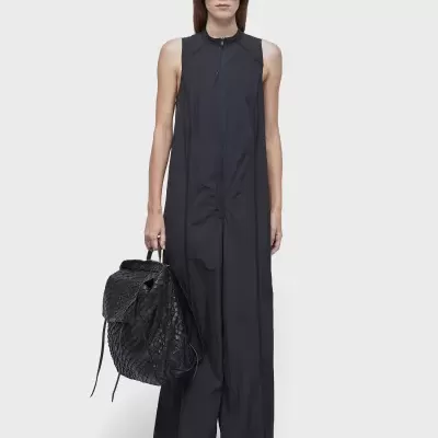 Mulher com vestido preto longo, mala preta e óculos escuros sobre fundo cinza claro