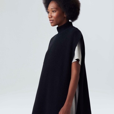 Mulher com capa preta de malha de gola alta por cima de roupa branca com mangas curtas em fundo cinza.