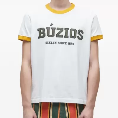 T-shirt branca com texto BÚZIOS OSKLEN SINCE 1989 e calças listradas verdes, amarelas e vermelhas