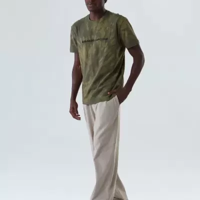 Homem com t-shirt verde camuflada, calças bege largas e sapatos claros em fundo branco neutro