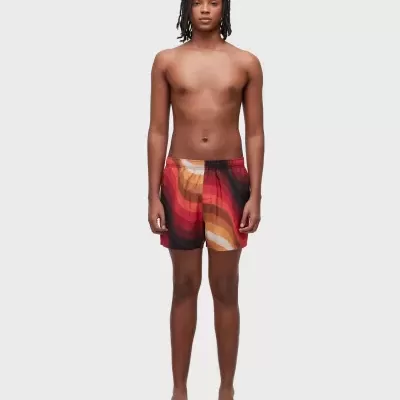 Homem com calções de banho multicoloridos e sandálias castanhas em fundo cinza claro.