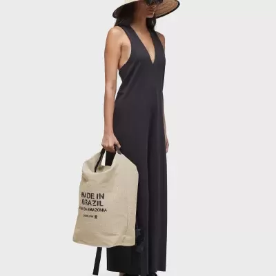 Mulher com macacão preto, chapéu de palha, óculos escuros, sandálias pretas e bolsa bege com texto preto MADE IN BRAZIL