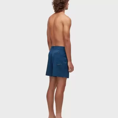 Homem com calções de banho azuis vistos de costas, em fundo branco.