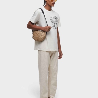 Homem com roupa casual clara, incluindo t-shirt branca estampada e calças bege, com mala trançada bege e óculos de sol amarelos.