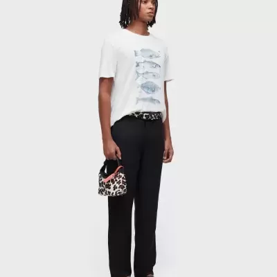 Pessoa vestindo t-shirt branca com peixes, calças pretas, cinto e mala animal print, sandálias pretas.
