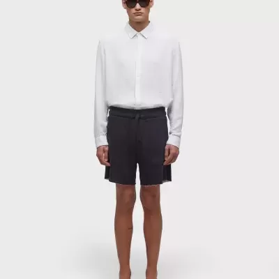 Modelo de pé com camisa branca, calções pretos e sandálias pretas em fundo branco