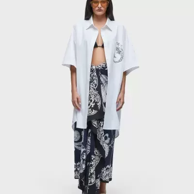 Mulher com camisa branca oversize, saia/calças largas pretas com padrão branco e óculos laranja