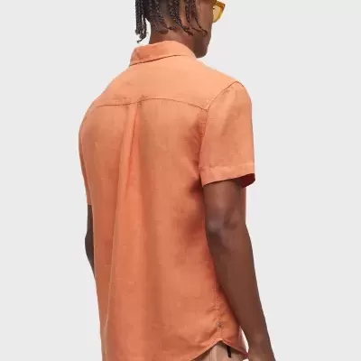 Camisa laranja de manga curta vista de costas, sem padrões, usada por modelo com tranças e óculos amarelos.