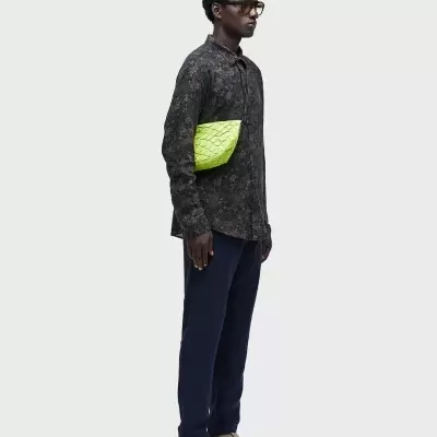 Homem com camisa preta, calças azuis escuras, sandálias bege e carteira verde fluorescente com textura acolchoada