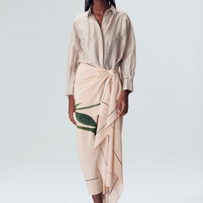 Mulher com camisa bege texturizada e saia longa bege com estampas verdes e linhas pretas, com ténis brancos.