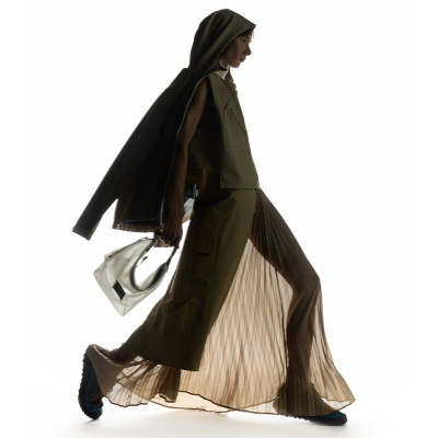Modelo com vestido plissado castanho e colete verde oliva, capa com capuz, mochila branca e sapatos com detalhe azul.