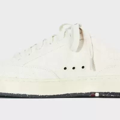 Tênis branco de tecido com textura cruzada e sola preta com detalhes.