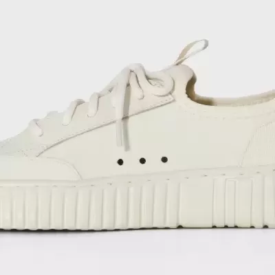 Ténis brancos com sola grossa e cadarços brancos em fundo neutro.