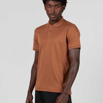 Homem com polo castanha de manga curta e calças pretas em fundo cinzento