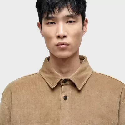 Homem jovem com camisa bege de tecido grosso com bolsos frontais e botões escuros.