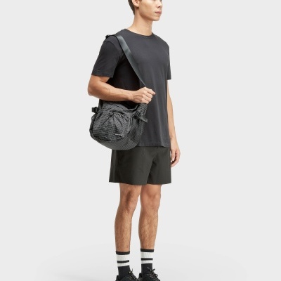 Homem vestido com t-shirt preta, calções, meias brancas com riscas pretas e sapatos pretos, a usar bolsa preta de ombro com padrão xadrez e texto OSKLEN.