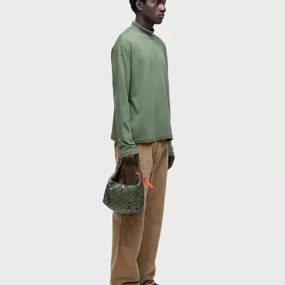 Homem com camisola verde, calças castanhas, sapatos pretos e mala verde com fita laranja em fundo branco