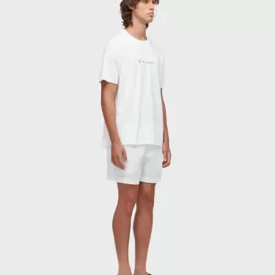 Homem com t-shirt branca, calções brancos e sandálias pretas em fundo cinzento