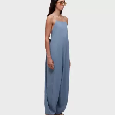 Mulher com macacão azul claro de tecido fluido e alças finas, usando óculos de sol castanhos e sapatos beige.