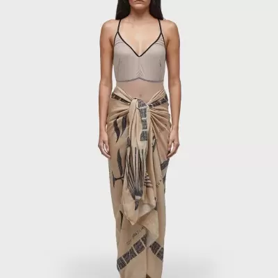 Mulher a usar top cinza claro e saia pareo bege com padrões pretos amarrada à frente