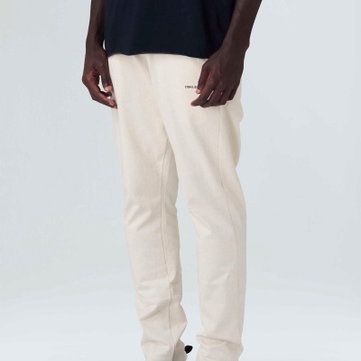 Calças bege claras com texto preto no lado esquerdo, camisola preta e ténis pretos, fundo cinzento claro