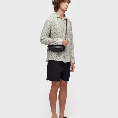 Homem com camisa de manga comprida estampada, calções pretos, mochila preta a tiracolo e chinelos pretos