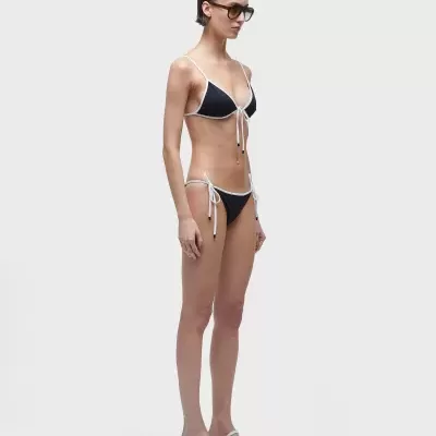 Biquíni preto e branco com top triangular e calças com laços laterais, usado por mulher com óculos escuros e sandálias brancas em fundo cinzento claro.