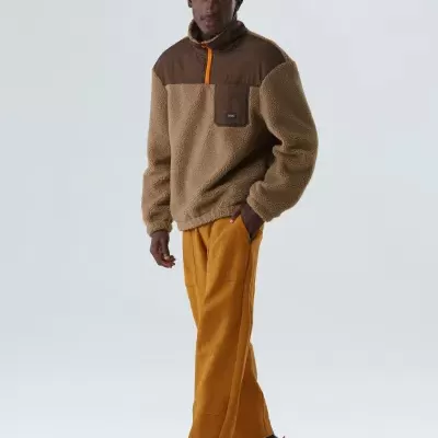 Homem com casaco bege e castanho, calças castanhas e ténis brancos em fundo branco