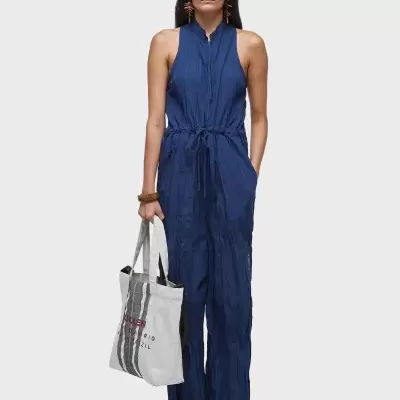 Mulher com macacão azul escuro, óculos escuros, pulseira castanha, sapatilhas brancas e tote bag branca com riscas pretas e texto.