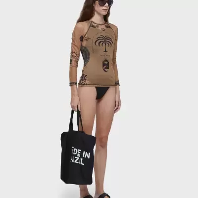 Mulher com camisa manga comprida castanha estampada, calção de banho preto e tote bag preta com texto branco