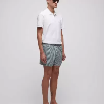 Homem com camisa polo branca, calções de banho riscas verdes e brancas, chinelos pretos e óculos de sol em fundo cinzento.