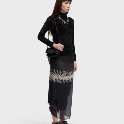 Mulher vestindo blusa preta de veludo, saia preta com estampa, colar dourado e mala preta ao ombro.