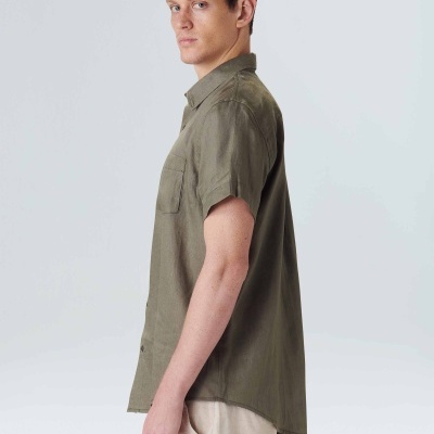 Homem de perfil com camisa verde-oliva de manga curta e bolso, calças claras, fundo branco