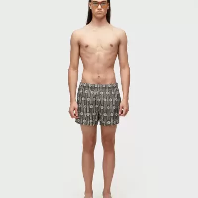 Homem com calção de banho preto com padrão branco, óculos de sol e chinelos brancos.