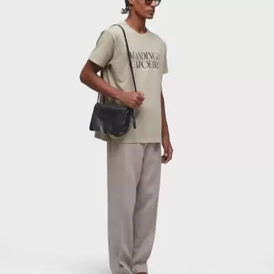 Homem com t-shirt bege com texto, calças cinza, chinelos castanhos, óculos escuros e bolsa preta