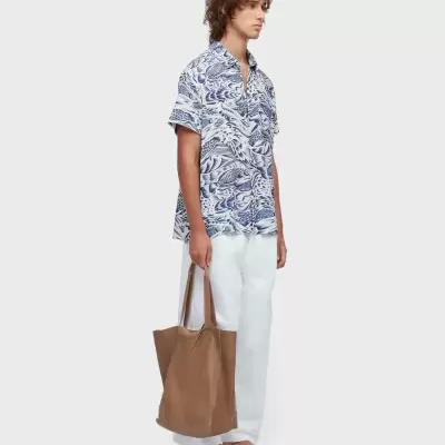 Homem com camisa azul e branca, calças brancas, chinelos bege e mala castanha