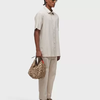 Homem com camisa e calças bege, segurando bolsa trançada e usando óculos amarelos