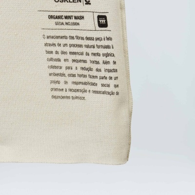 Etiqueta de tecido cru com texto preto em português, marca OSKLEN e descrição de amaciamento de fibras e projeto ambiental