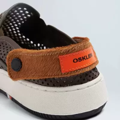 Calçado estilo sapatilha castanho com sola branca e preta, tira traseira castanha com etiqueta laranja OSKLEN