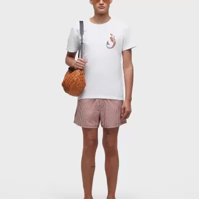 Homem de t-shirt branca com estampa de lagartixa, calções riscas rosa e brancas, sandálias castanhas, óculos laranja e mala de malha laranja