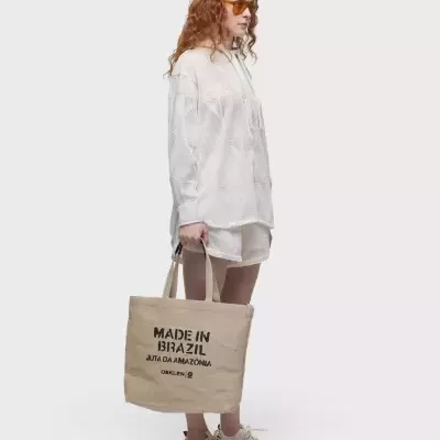 Mulher com hoodie branco e sacola bege com texto 'MADE IN BRAZIL JUTA DA AMAZÔNIA OAKLEY'