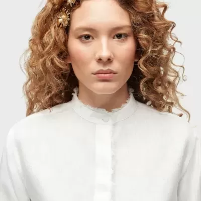 Blusa branca com gola redonda e detalhes irregulares, mulher com cabelo ruivo cacheado e presilhas de flor dourada