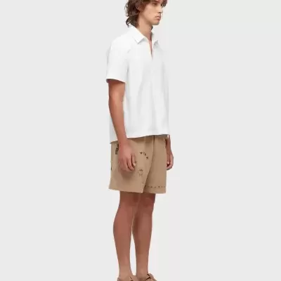 Homem com camisa branca, calções beges com desenhos e sandálias beges.