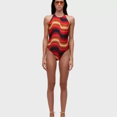 Fato de banho com padrão ondulado em tons quentes, usado por mulher com óculos de sol laranja e sapatos de corda bege num fundo cinza.