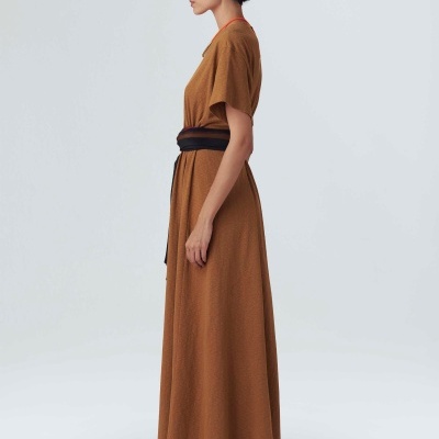 Vestido comprido castanho com cinto preto usado por modelo em fundo neutro.