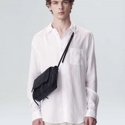 Homem com camisa branca e bolsa preta tiracolo em fundo cinzento claro.