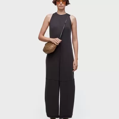 Mulher com vestido preto largo e sem mangas, mala castanha entrelaçada, ténis bege e óculos de sol com lentes âmbar.