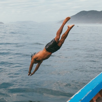 Rapaz a mergulhar no mar perto de um barco colorido com montanhas ao fundo