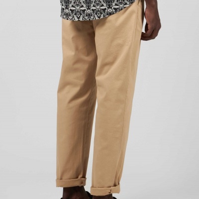 Pessoa usando calças bege, camisa preta e branca com padrão e sapatilhas pretas em fundo cinzento